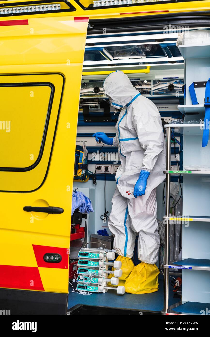 Side view of anonymous doctor in protective suit standing in ambulance car near opened door with equipment and examining patient with virus infection Stock Photo