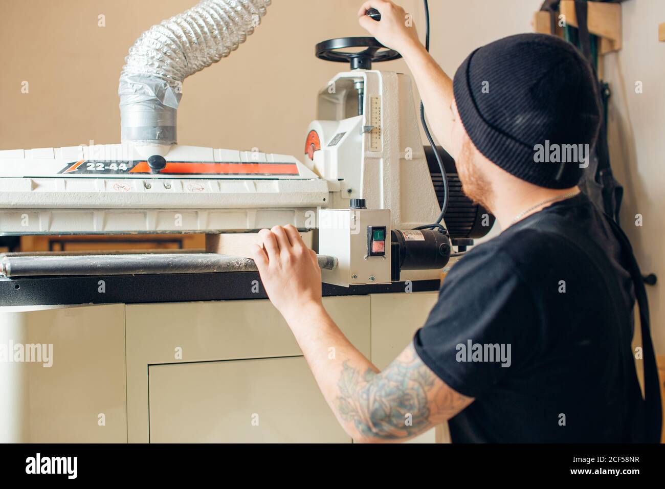 man in a gray cap carpenter builder in work clothes processing a tree with a milling machine in the workshop, around a lot of equipment, wooden boards Stock Photo