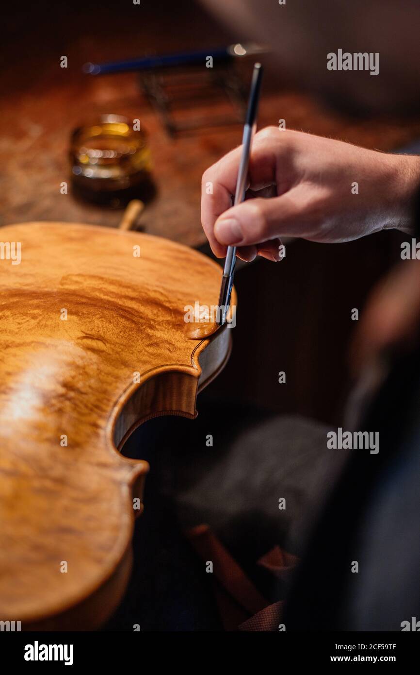 High angle of unrecognizable craftsman painting violin with thin brush while lacquering and coloring soundboard at table in workroom Stock Photo