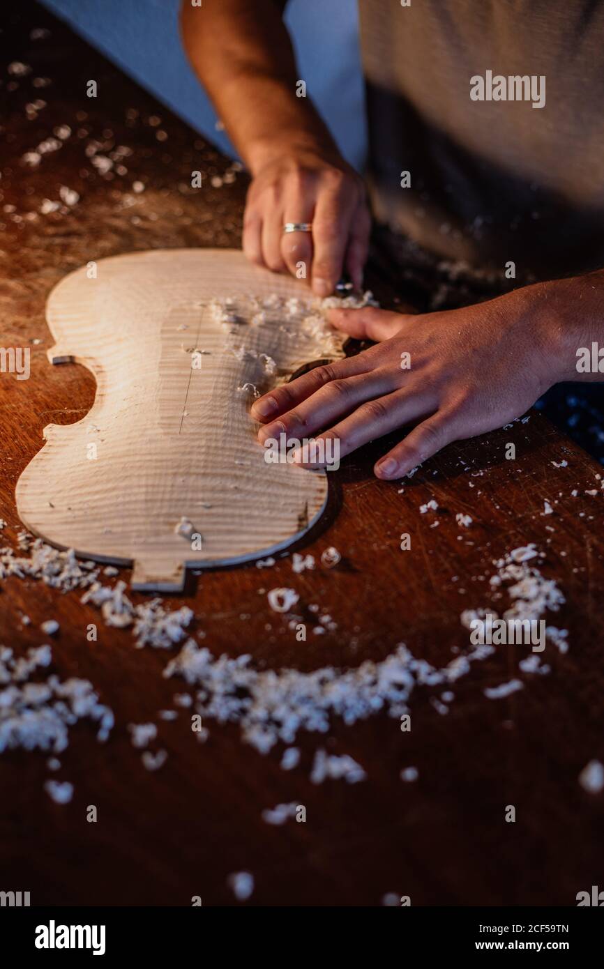 High angle of unrecognizable craftsman planing wooden violin soundboard using tool at table in work studio Stock Photo