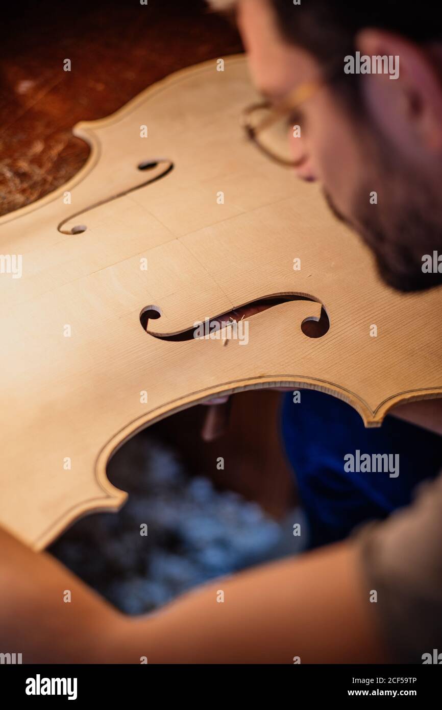 High angle side view of crop attentive artisan finishing cutting f holes in wooden violin soundboard at table in workroom Stock Photo