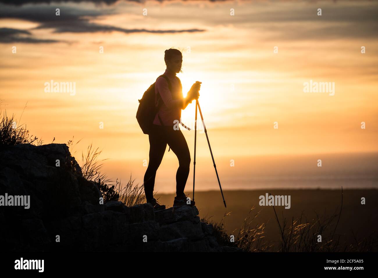 Silhouette of unrecognizable Woman standing with walking sticks on stony hill spreading arms wide in sunset Stock Photo