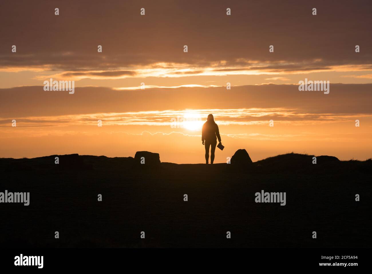 Woman silhouette with book standing at sunset opposite sun in rocky terrain in Wales Stock Photo