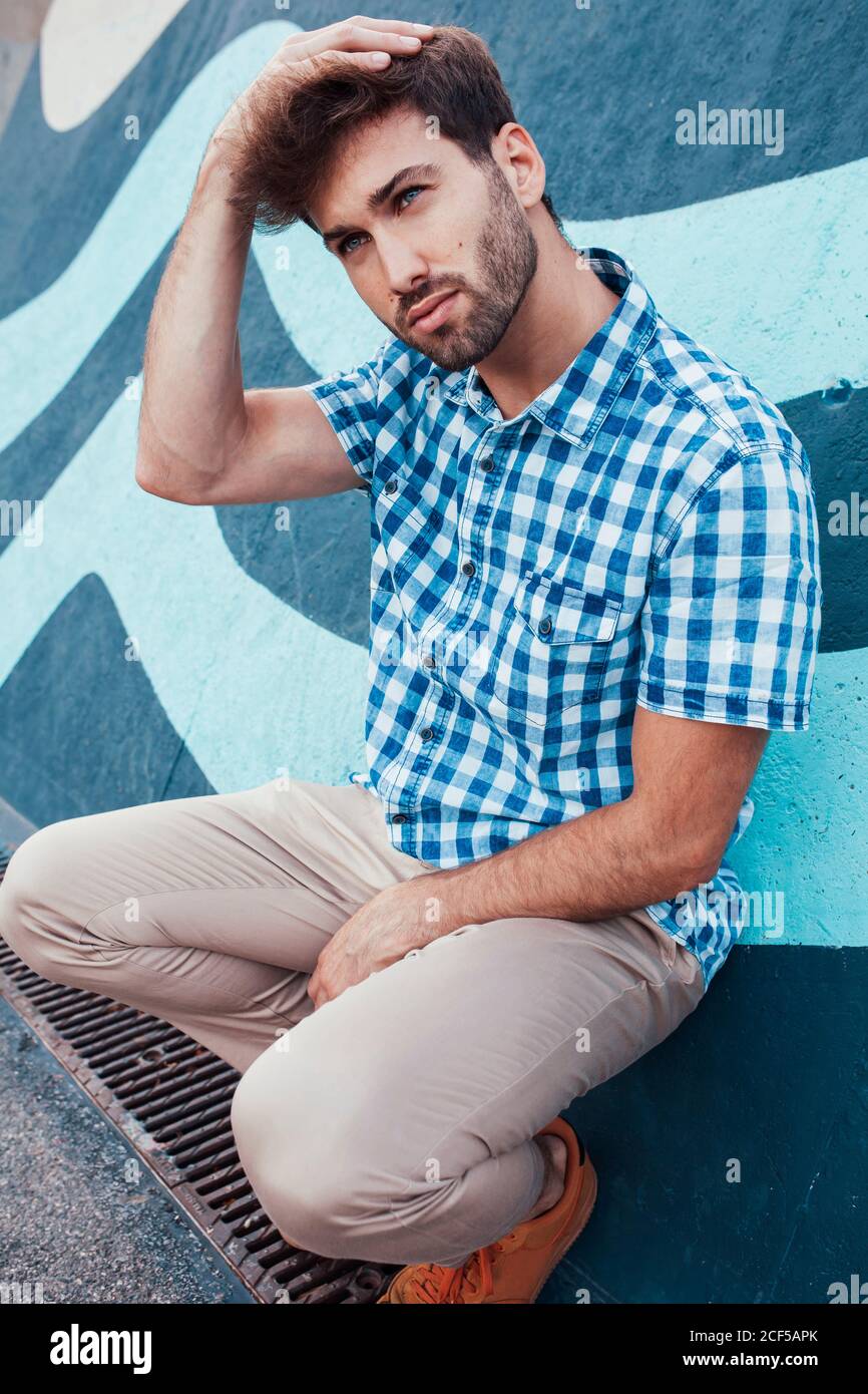 Cheerful young male in casual plaid shirt and sneakers squatting and looking away with painted wall on background Stock Photo