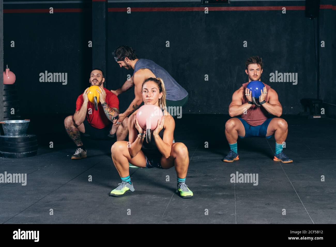 Company of muscular sportspeople squatting with kettlebells during intense workout under supervision of professional instructor Stock Photo