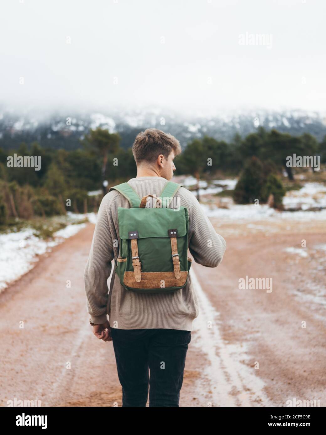 Back view of young male traveler in sweater with backpack walking on dirt rural road in cloudy winter day with green forest and snow on roadsides Stock Photo