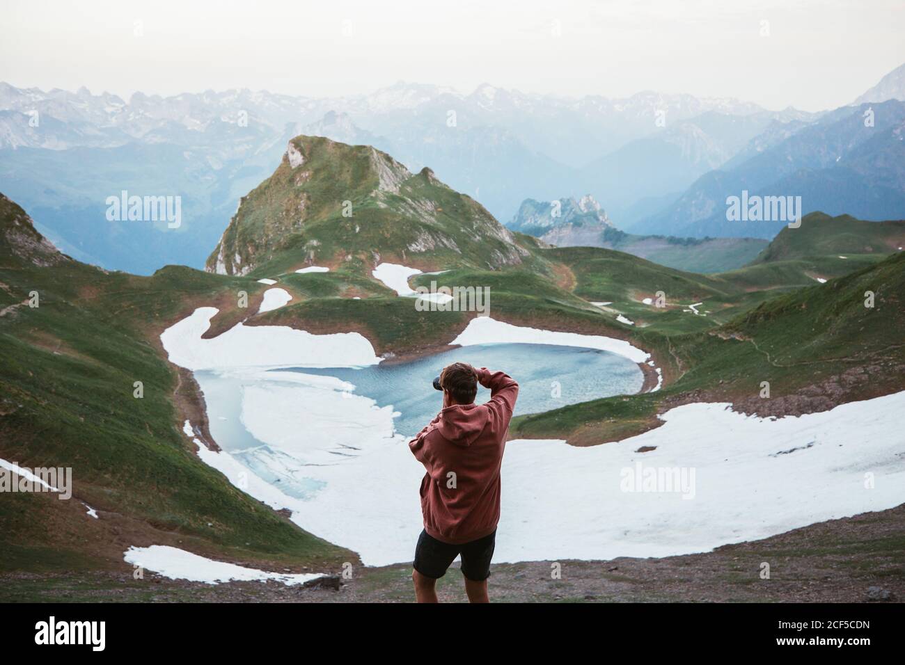 Back view of anonymous male tourist taking photo of mountain lake surrounded by snow and ridge in Pyrenees Stock Photo