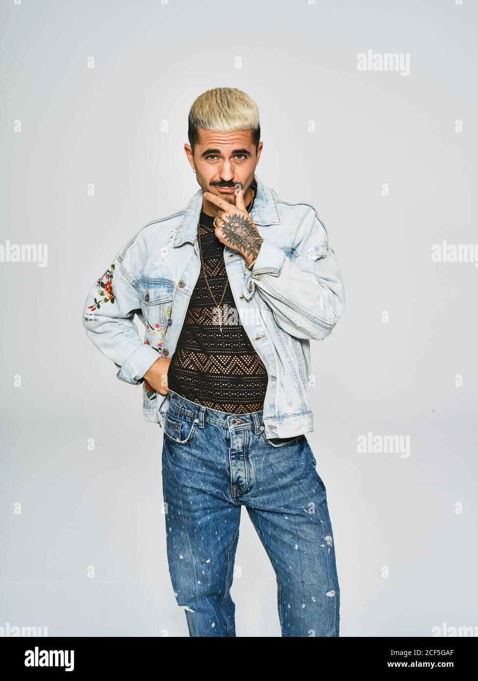Young ethnic man making grimace doubting face looking at camera wearing trendy denim jacket with floral pattern while standing against gray background Stock Photo