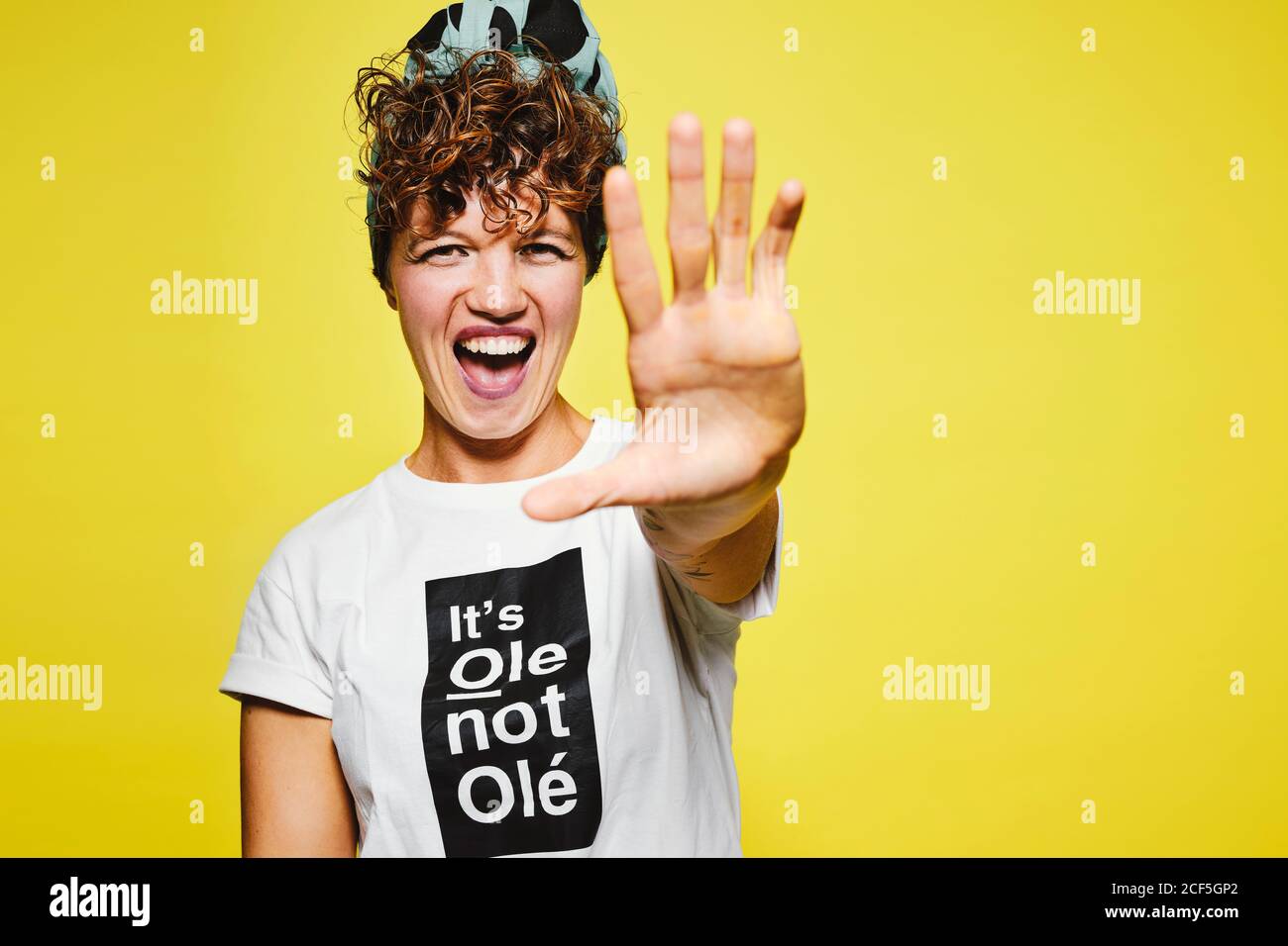 Positive adult female in white t shirt and trendy headscarf looking at camera with opened mouth and stretching out hand against yellow background Stock Photo
