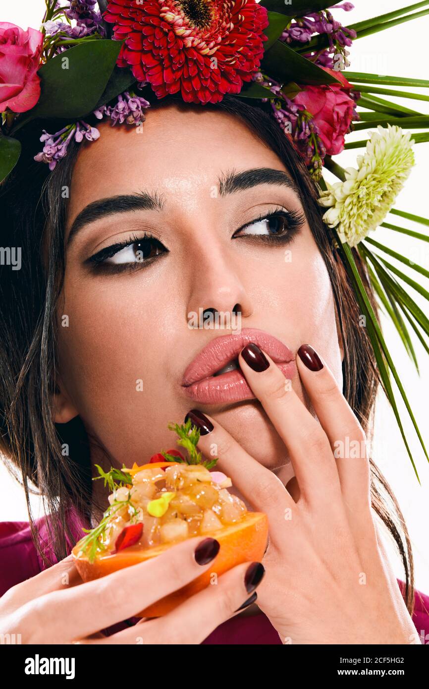 Young gorgeous Woman with wreath of colorful flowers on head and finger on lip looking away while holding dish in orange zest isolated on white background Stock Photo