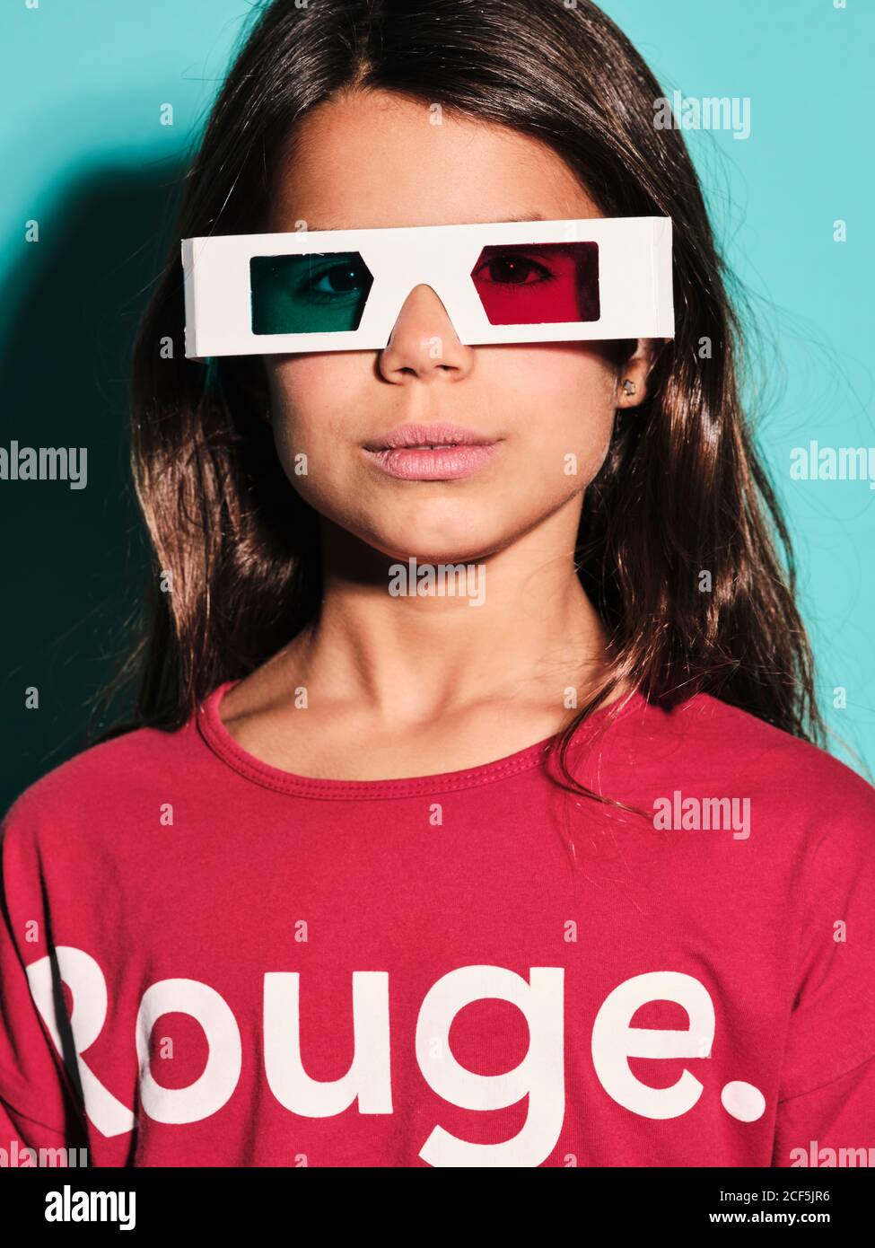 Portrait of happy preteen girl in white plastic stereoscopic glasses with red and blue lens smiling at camera while watching 3d graphic film with pleasure against turquoise background in modern studio Stock Photo