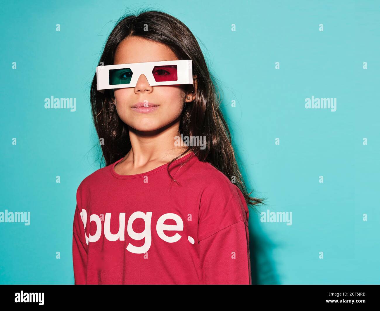Happy preteen girl in white plastic stereoscopic glasses with red and blue lens smiling at camera while watching 3d graphic film with pleasure against turquoise background in modern studio Stock Photo