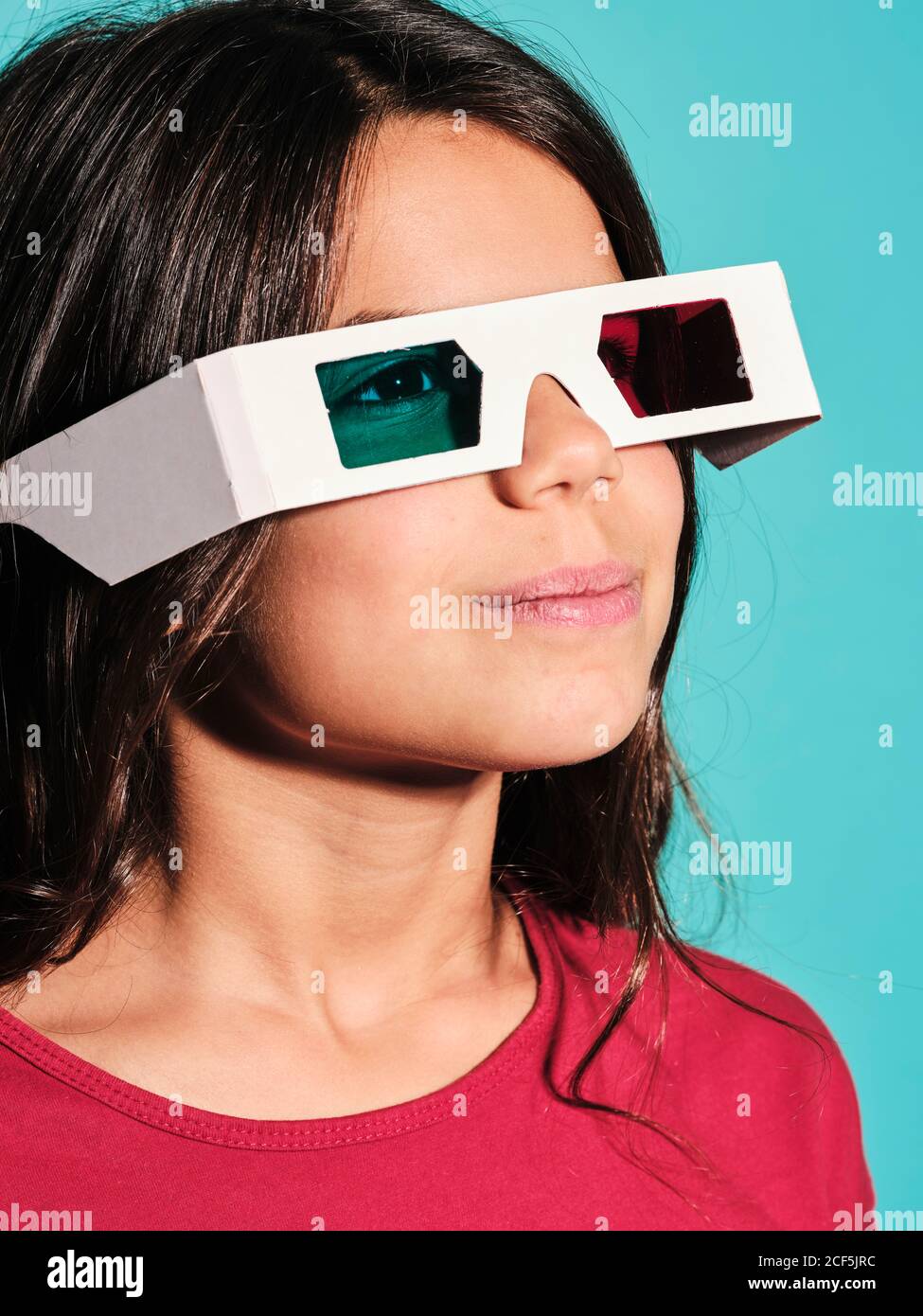 Portrait of happy preteen girl in white plastic stereoscopic glasses with red and blue lens smiling away while watching 3d graphic film with pleasure against turquoise background in modern studio Stock Photo