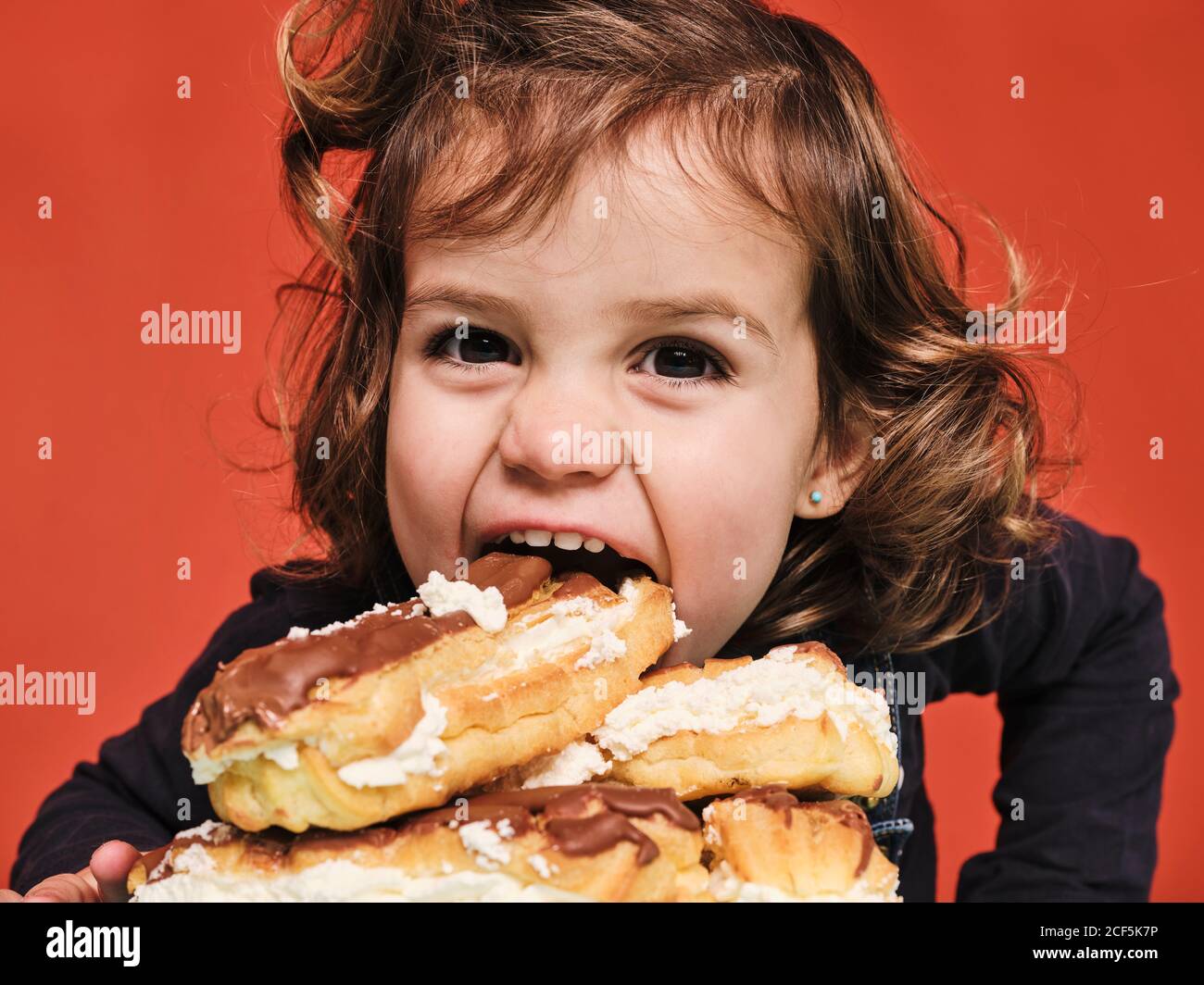 Closeup portrait of cheerful little girl enjoying sweet eclairs with chocolate while looking at camera against red background Stock Photo