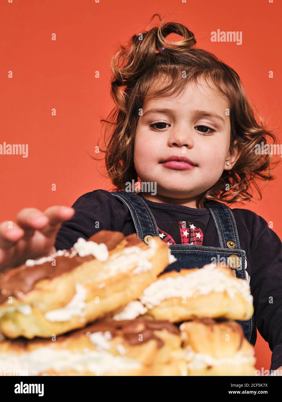 Happy little girl enjoying sweet eclairs with chocolate while sitting at table against red background Stock Photo