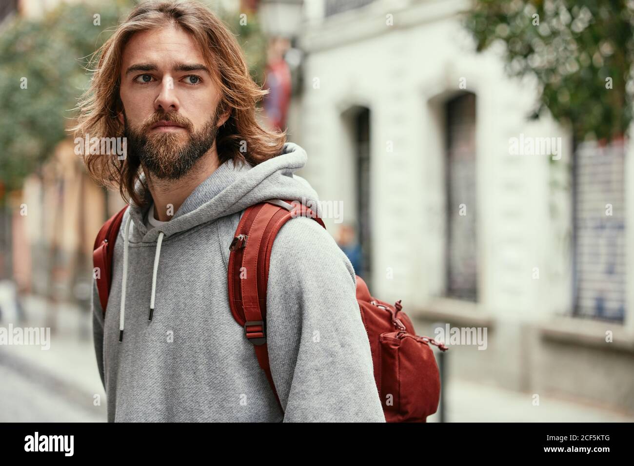 Young bearded handsome man in grey hoodie with backpack seriously walking outside Stock Photo