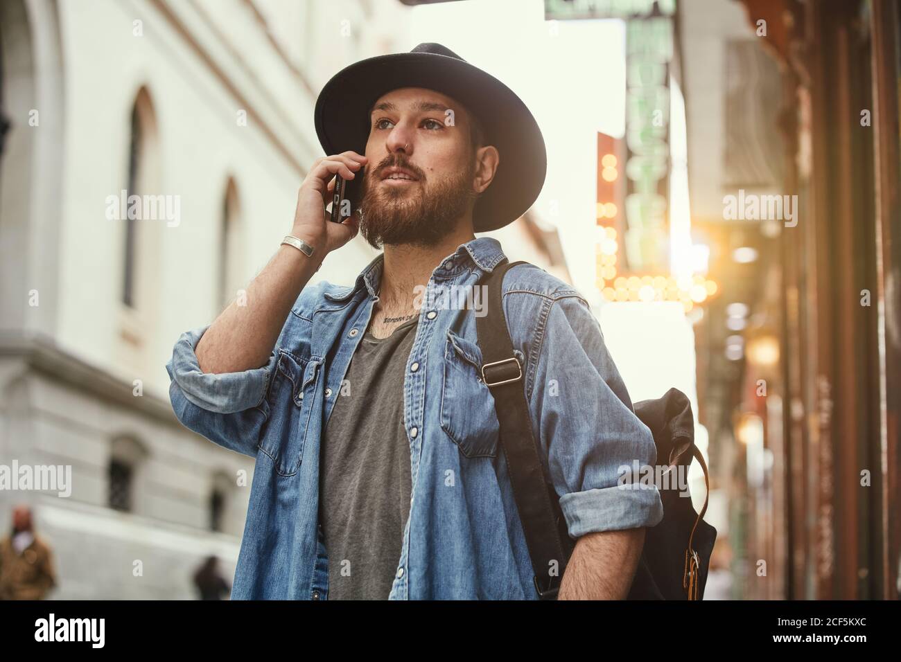 Young bearded handsome man in black hat and denim jacket merrily talking on mobile phone in street Stock Photo