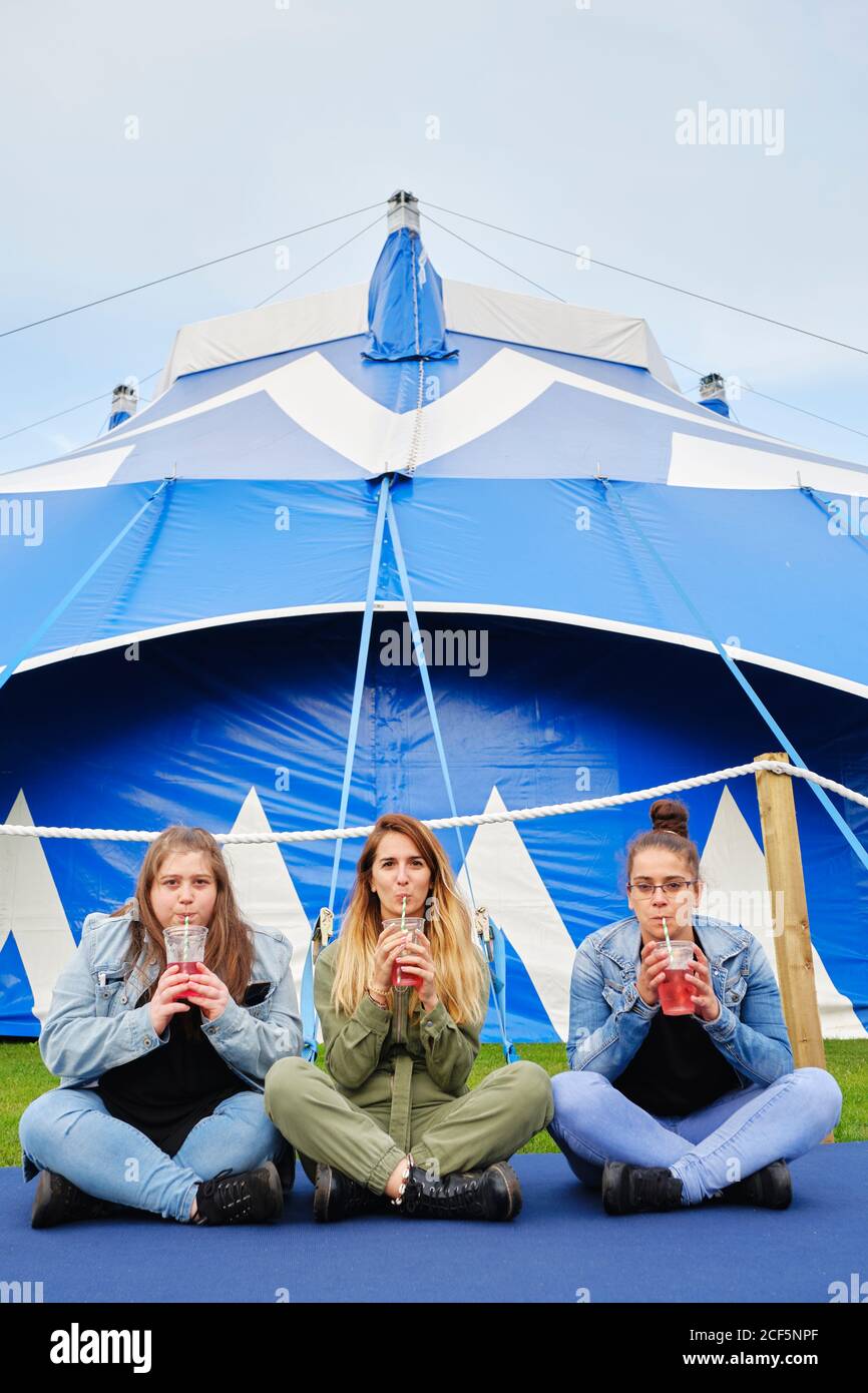 Cheerful young women in casual clothes drinking red beverage through straw while sitting on blue mat near fenced tent Stock Photo