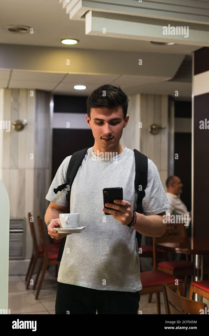 Young man with backpack carrying cup of hot beverage and browsing smartphone while standing in cafeteria Stock Photo