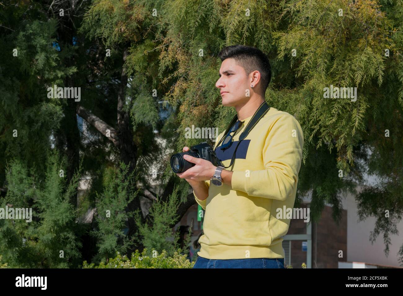 Low angle of black haired thoughtful male photographer in casual wear standing with photo camera and looking away in green park Stock Photo