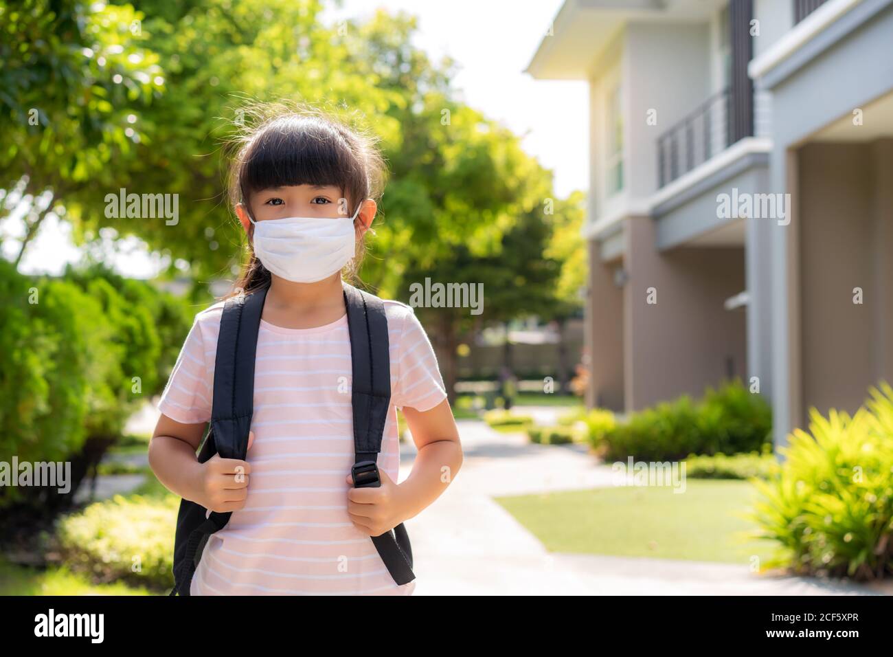 Portrait of cute Asian primary school girl in medical mask in front of home during COVID-19 pandemic. The morning school routine for day in the life g Stock Photo