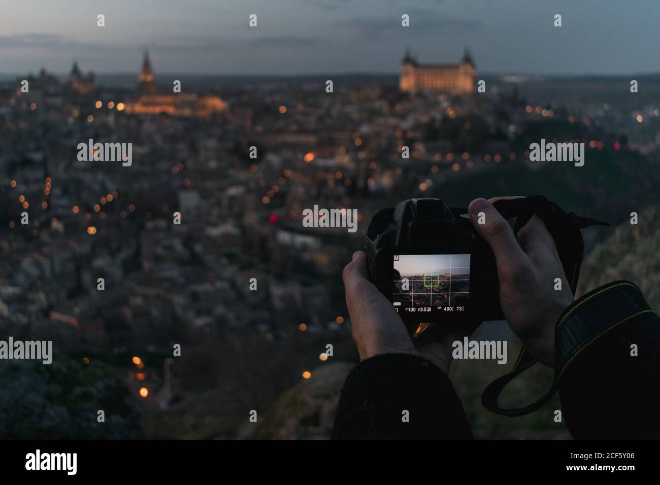 Crop hands of male photographer with camera standing on hill and taking photo of old Spanish city Toledo with castles in twilight Stock Photo