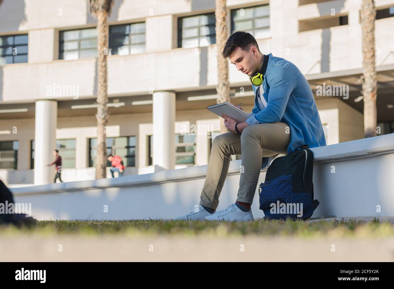 Side view of thoughtful smart male student in bright headphones studying in university square and writing in note book sitting on fence Stock Photo