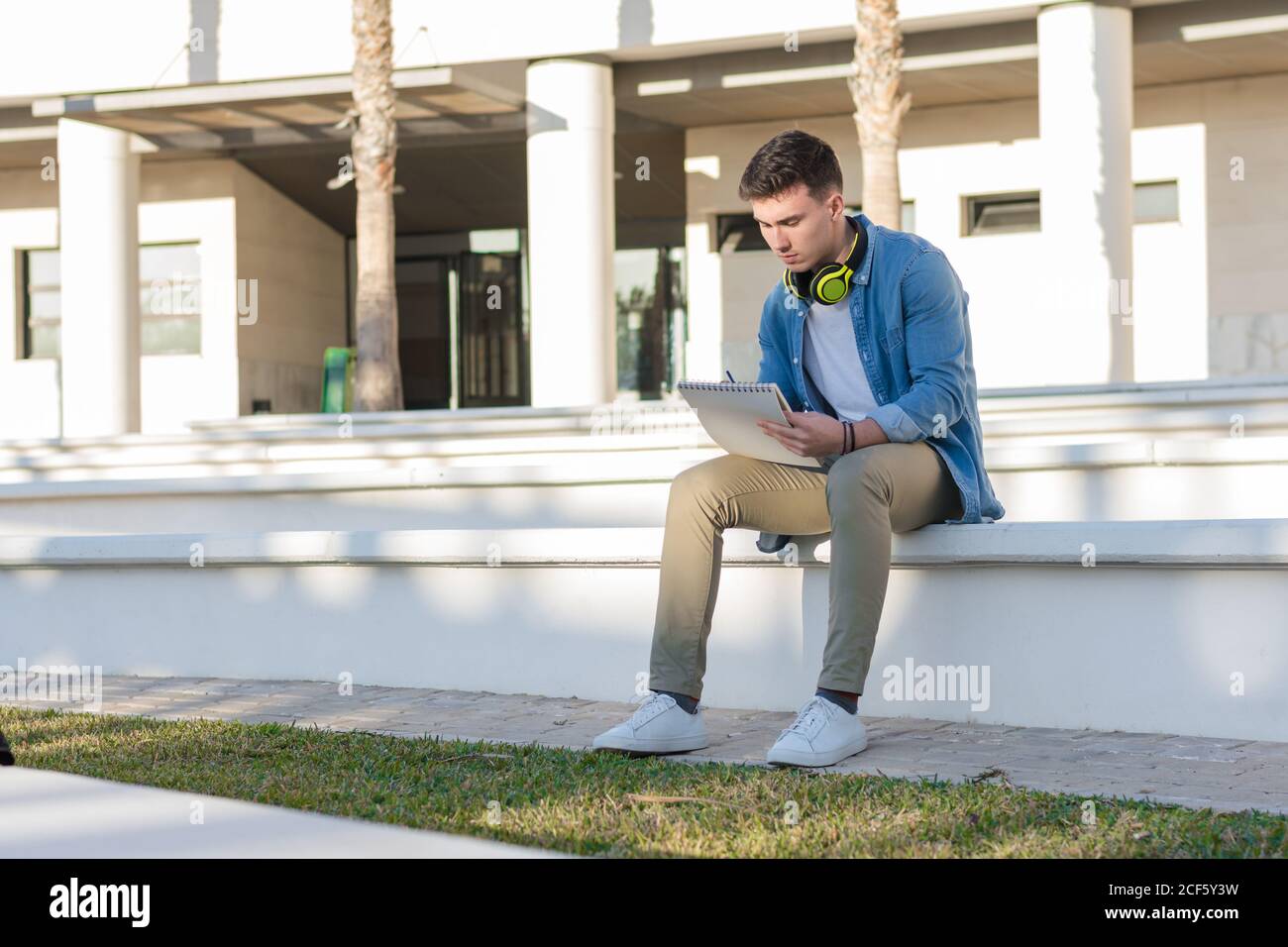 Thoughtful smart male student in bright headphones studying in university square and writing in note book sitting on fence Stock Photo