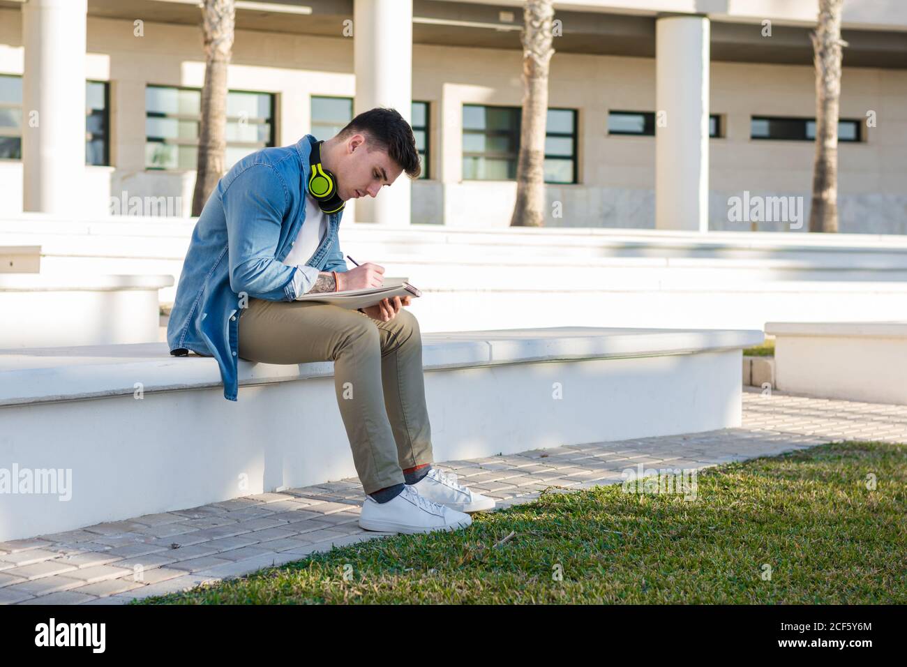 Side view of thoughtful smart male student in bright headphones studying in university square and writing in note book sitting on fence Stock Photo