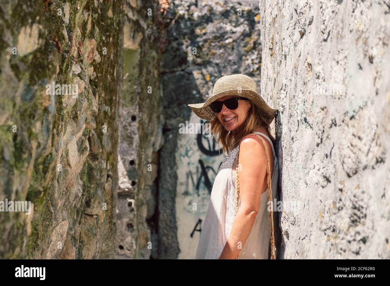 Tanned stylish Woman in hat, sunglasses and light costume enjoying sightseeing leaning in ancient stone wall looking at camera Stock Photo