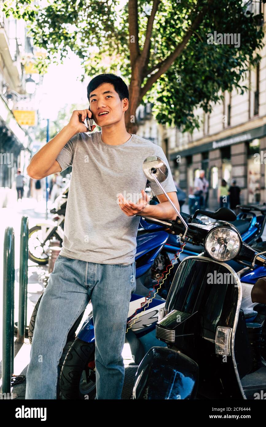 Positive casual ethnic man speaking on smartphone and gesturing while standing by motorcycle on sunny street Stock Photo