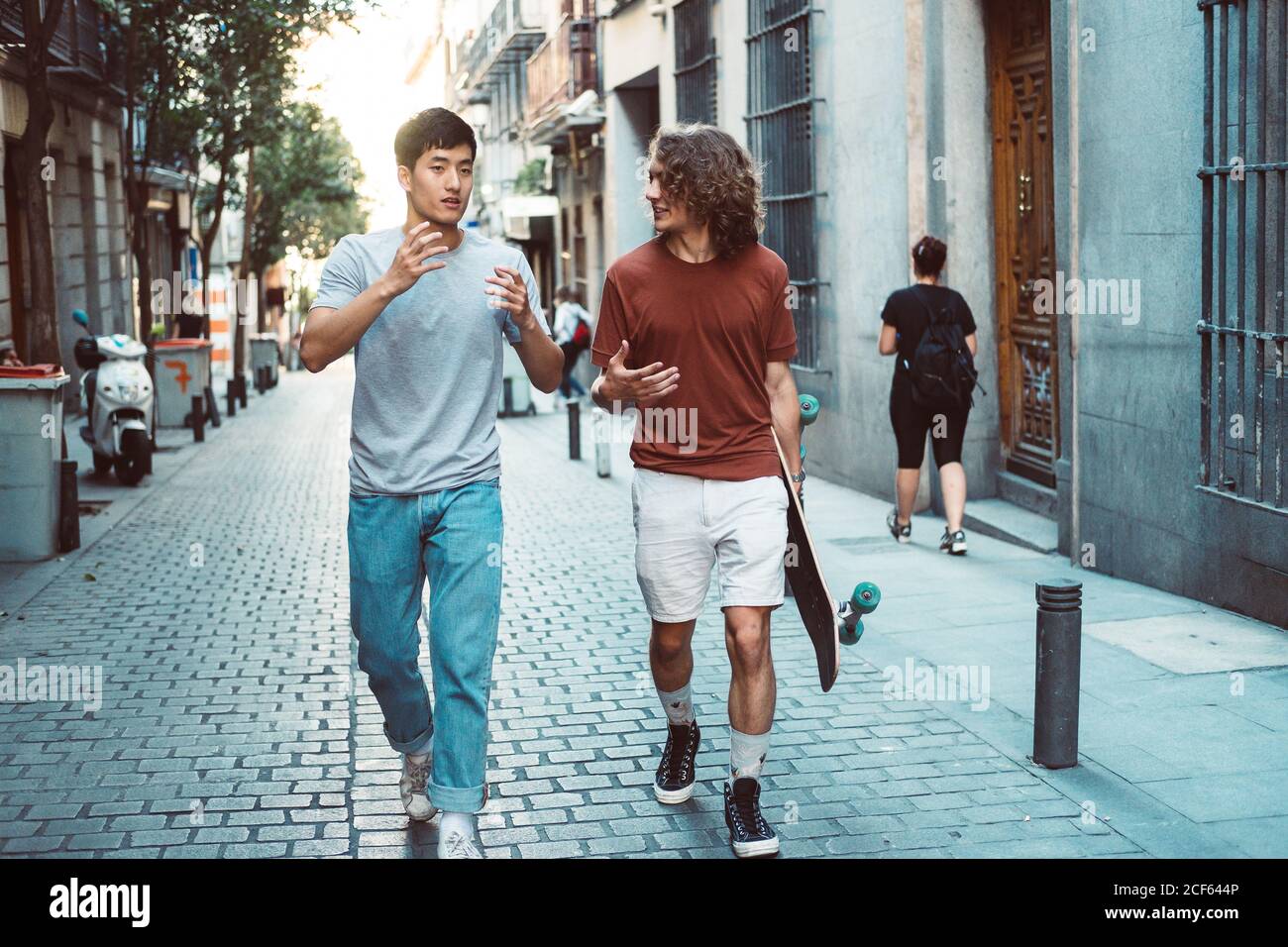 Carefree interested multiethnic men in casual clothes with longboard gesturing and talking while strolling along city street Stock Photo