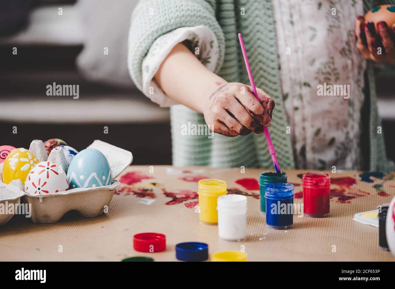 Crop kid with dirty hands in paints coloring Easter eggs in container near bright colors on table Stock Photo