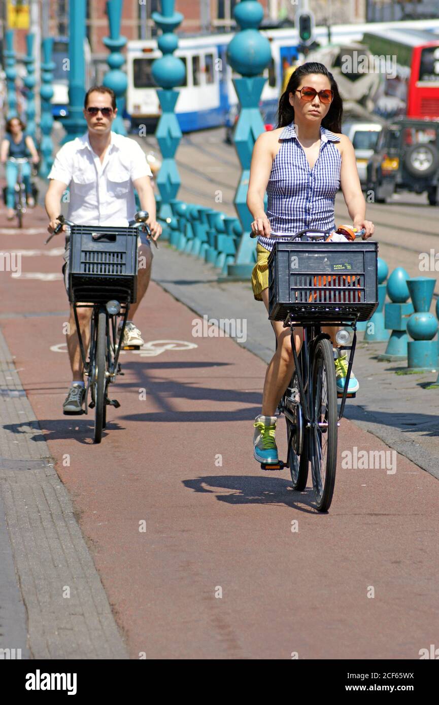 Two people ride two-wheel bicycles with baskets in a bike lane along Damrak in Amsterdam, Netherlands, Holland. Stock Photo