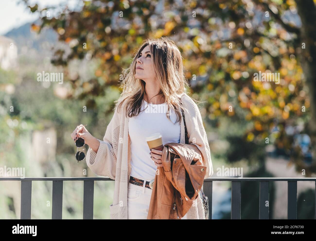 Attractive graceful woMan with coffee and jacket in hand looking along leaning on handhold in warm autumn day Stock Photo