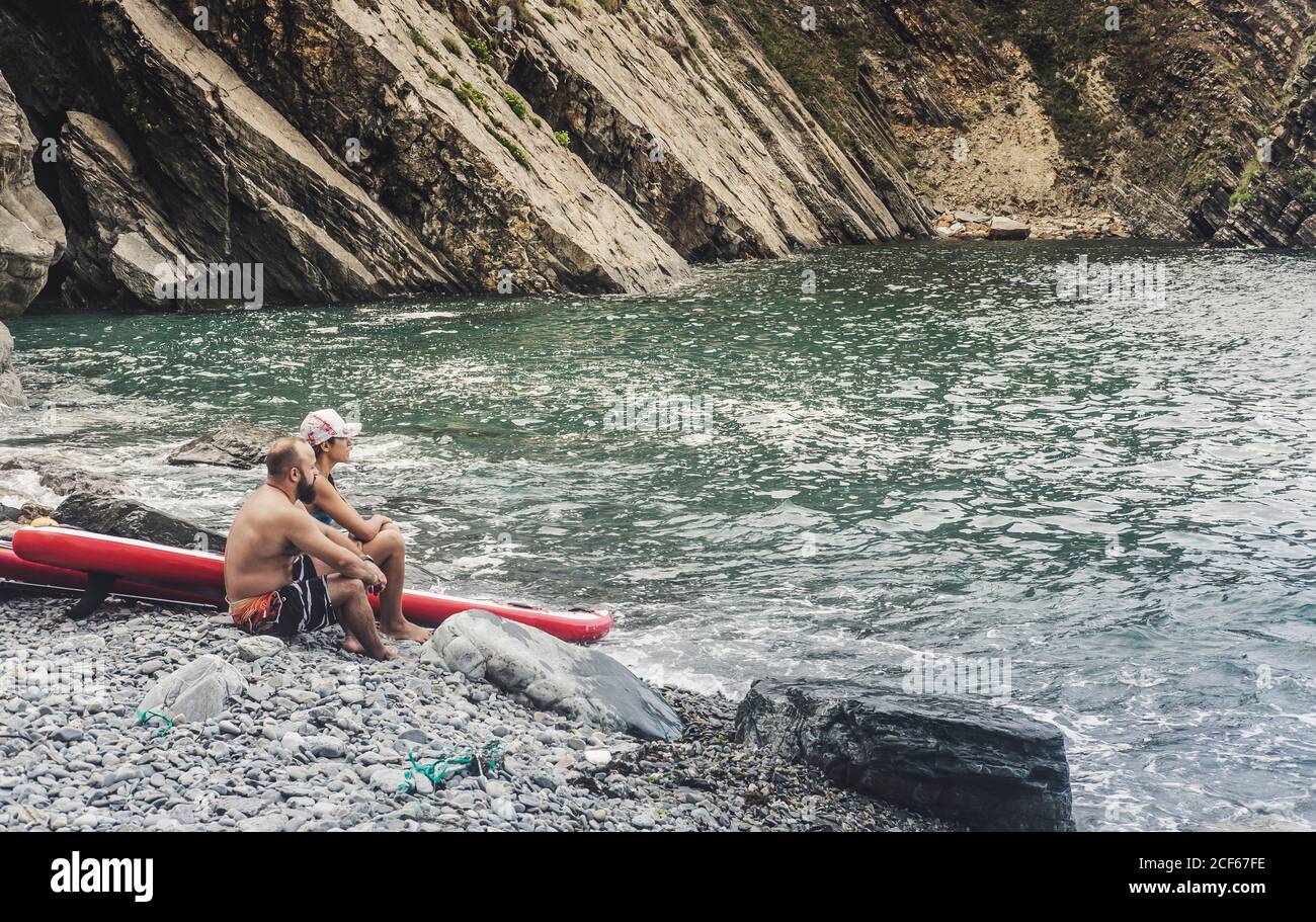 Side view of adult man and Woman sitting on rocky shore near paddleboards and admiring view of majestic sea Stock Photo