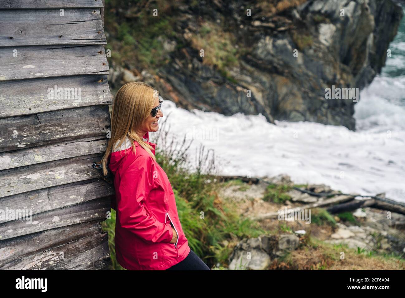 Woman in red raincoat leaning on house at shore looking at water Stock Photo