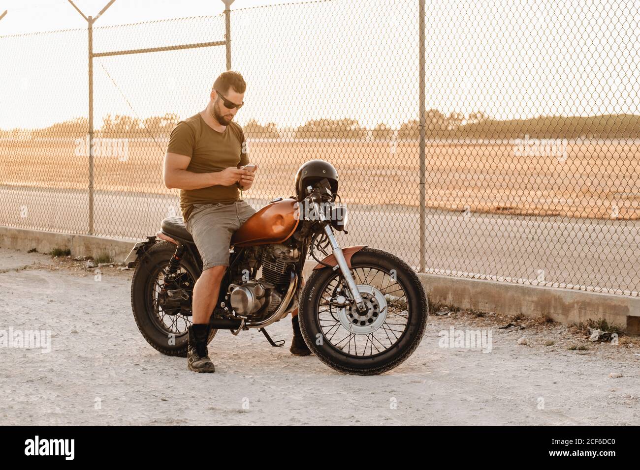 Wistful man in sunglasses having rest on motorcycle and using smartphone on field Stock Photo