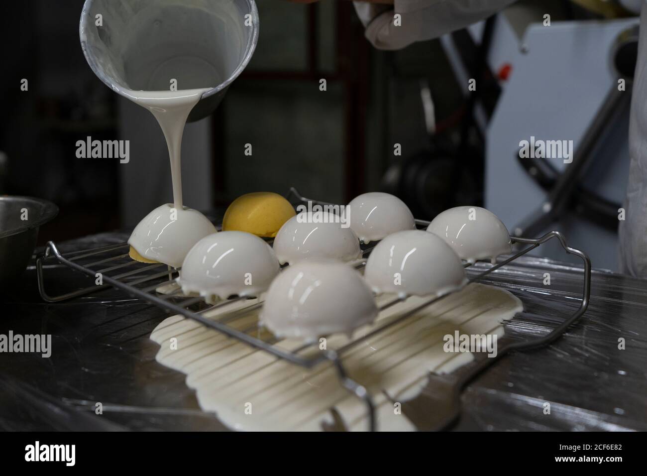 Crop person pouring white mirror glaze on tasty yellow mousse cakes arranged on metal grid in kitchen Stock Photo