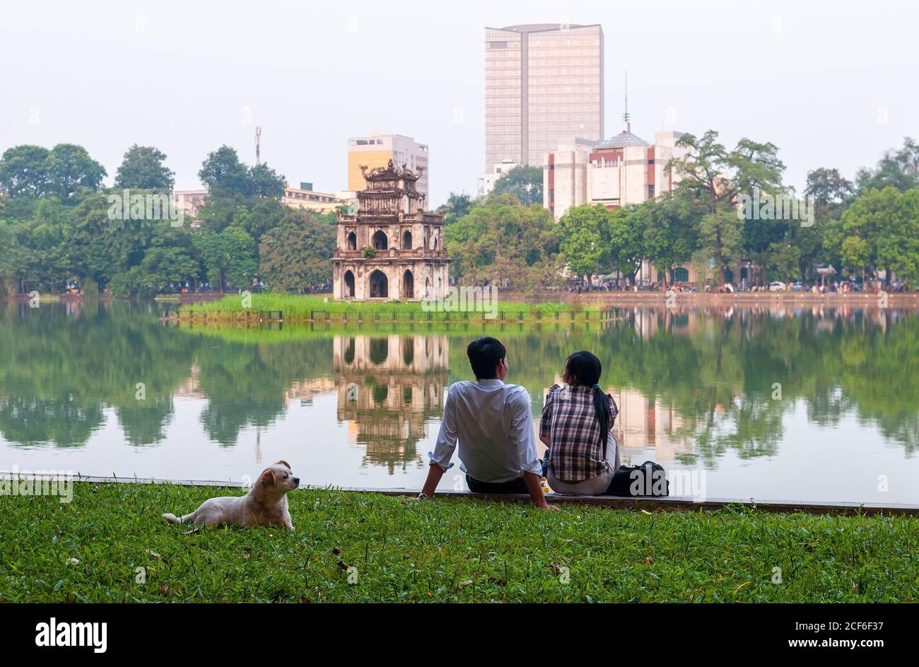 Friends talking along the Hoan Kiem lake with the turtle pagoda and modern buildings in the background in Hanoi, Vietnam. Stock Photo