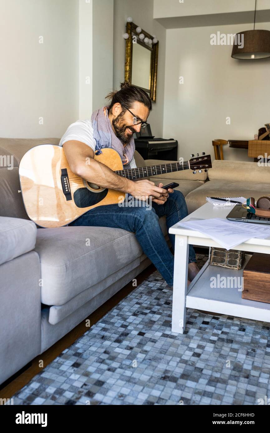 Cheerful man with guitar checking smartphone on sofa Stock Photo
