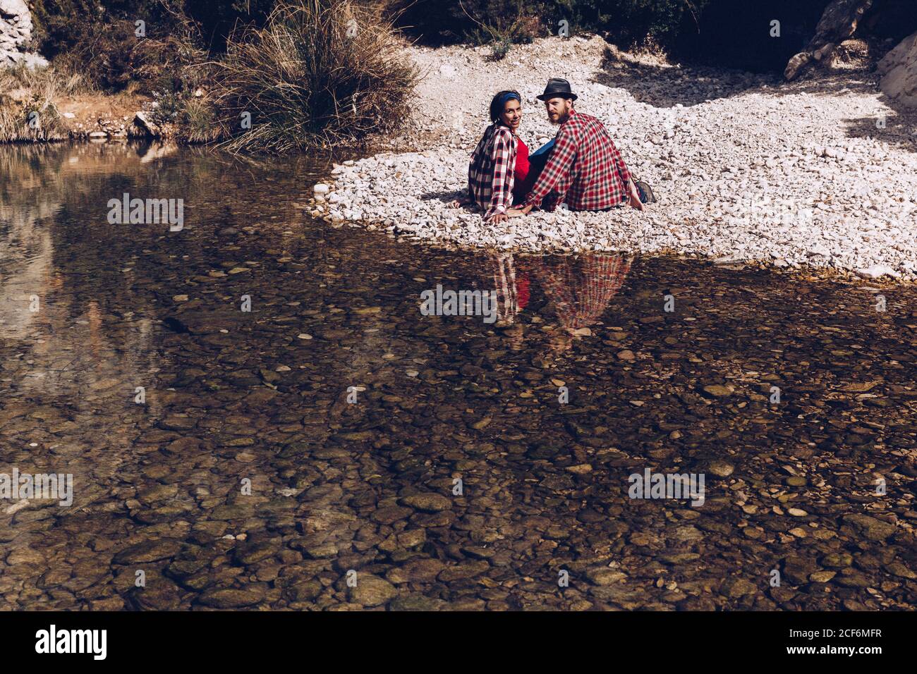 Side view of young couple sitting on rock coast of mountain river near cliff Stock Photo
