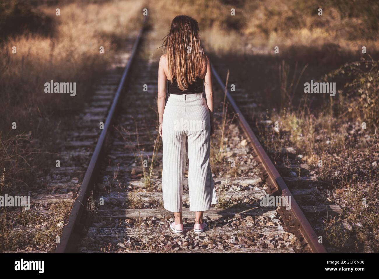 Back view of stylish Woman with long hair standing on railway ties through field of dry vegetation Stock Photo