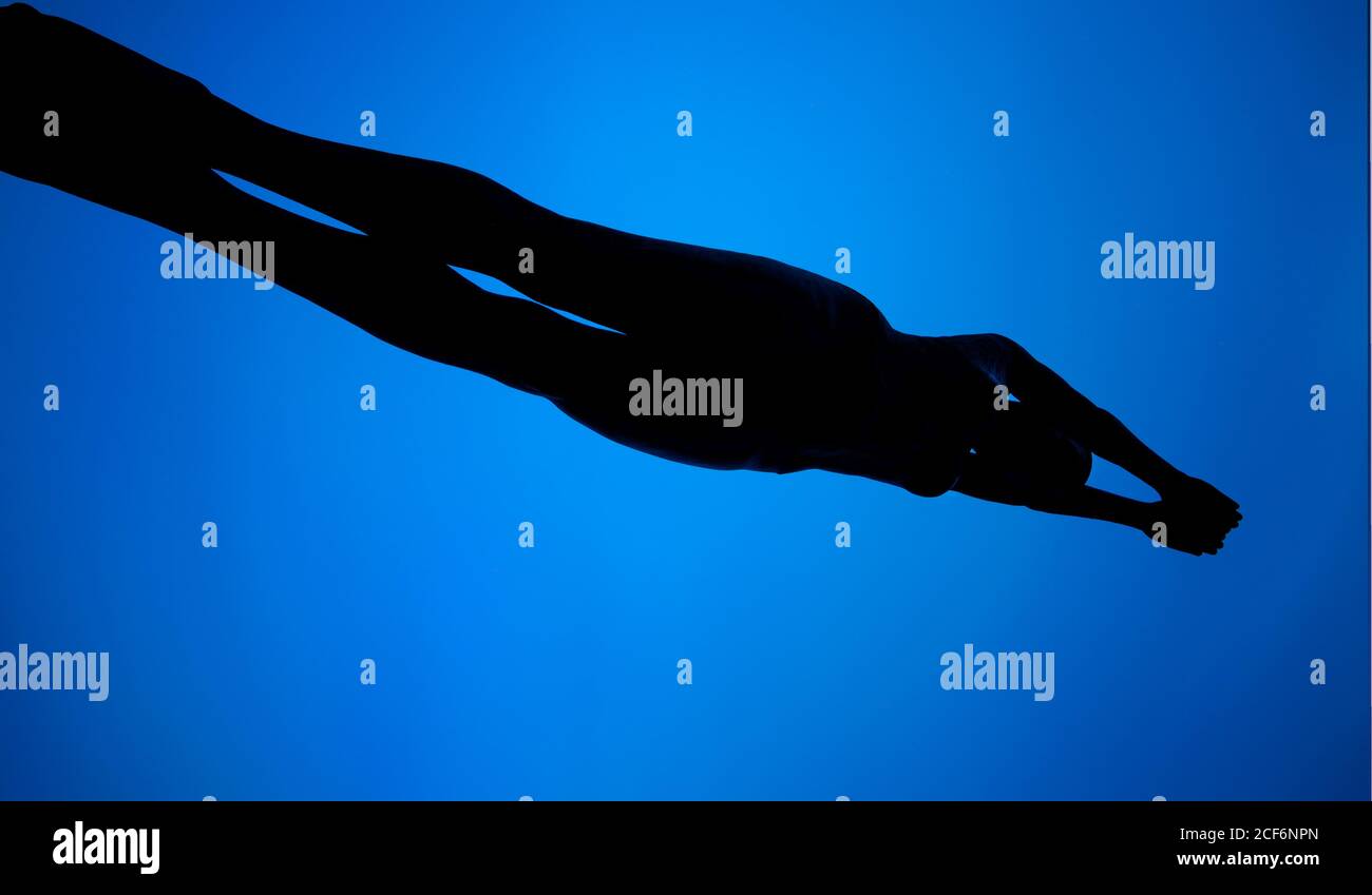 From below silhouette of slim anonymous female swimming under blue water Stock Photo