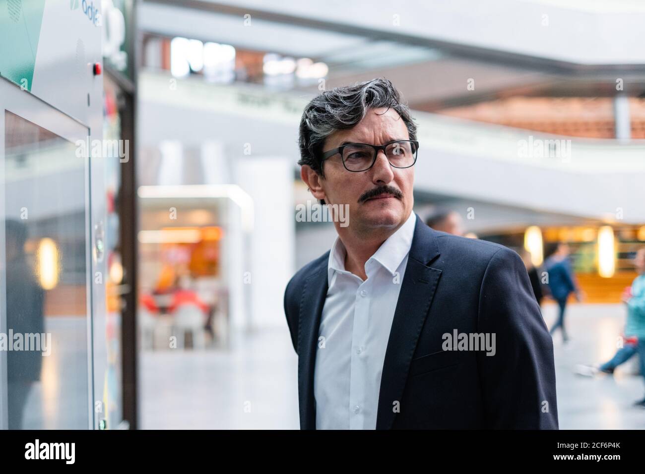 Bewildered handsome aged male entrepreneur in business style clothing and eyeglasses looking away on blurred background at shopping center Stock Photo