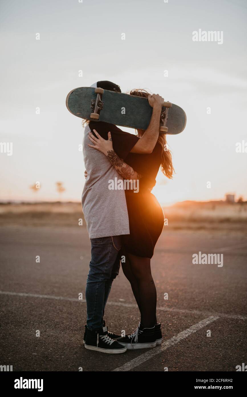 Side view of romantic teenage couple embracing and covering heads with skateboard while kissing in back lit of sunset on rural road Stock Photo