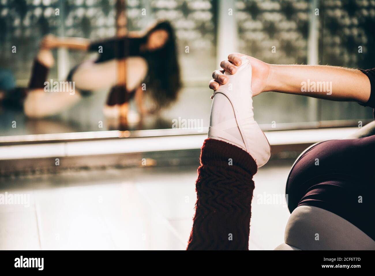 Crop shot of ballerina pulling leg to stretch muscles sitting on floor in ballet studio Stock