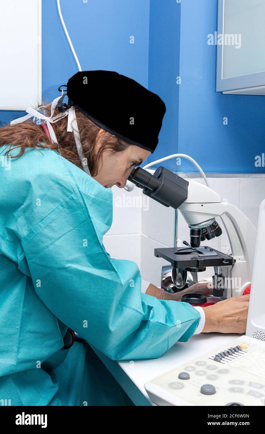 Side view of concentrated Woman in surgical gown and cap looking through microscope and doing research in lab Stock Photo