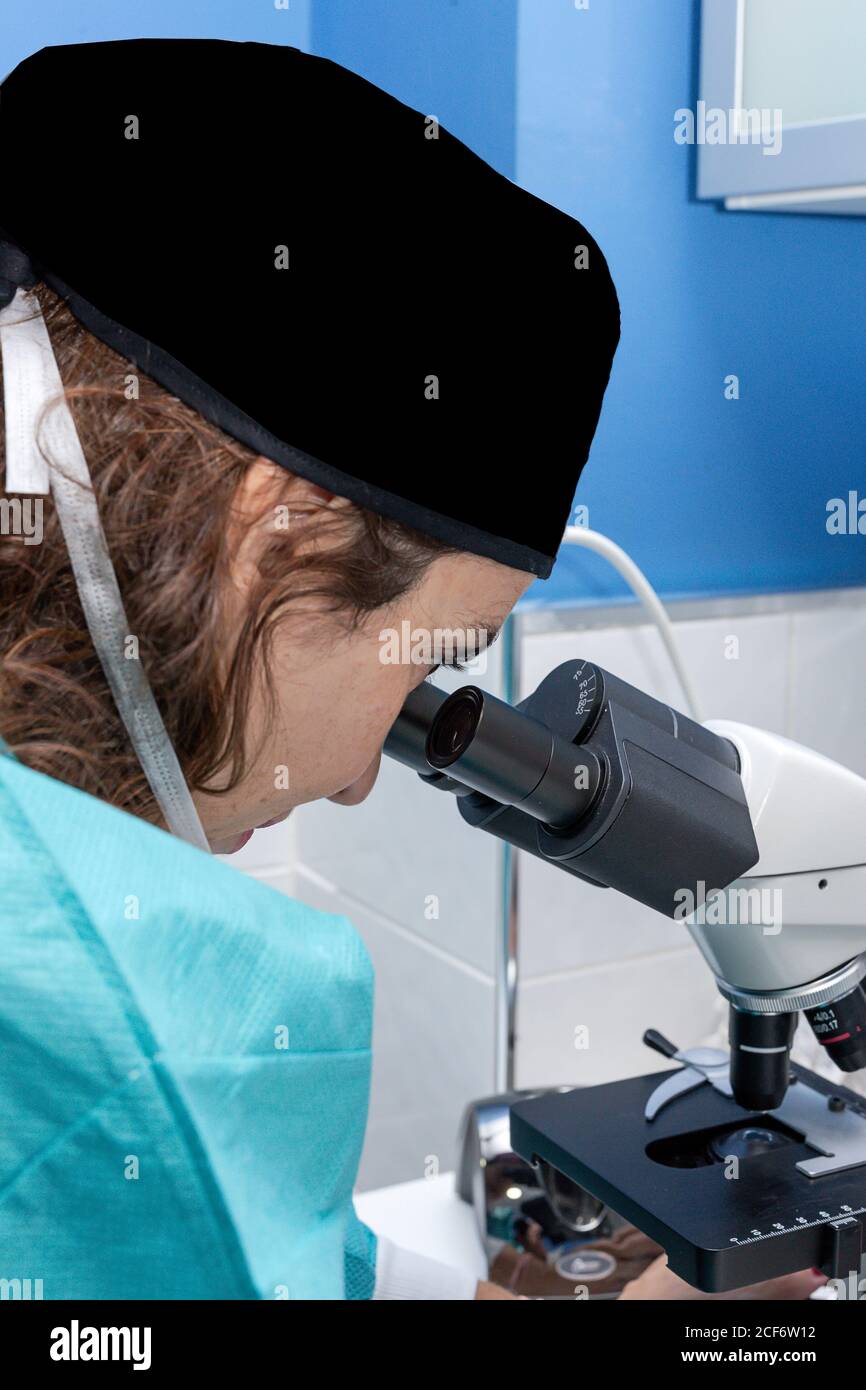 Side view of concentrated Woman in surgical gown and cap looking through microscope and doing research in lab Stock Photo