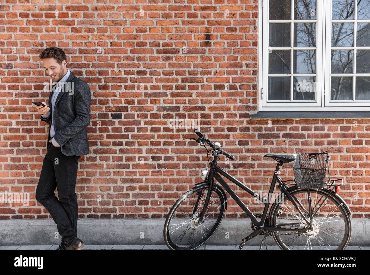 Young businessman going to work on bike commute using his mobile cellphone against city brick wall background. Happy business man bicycle commuter Stock Photo